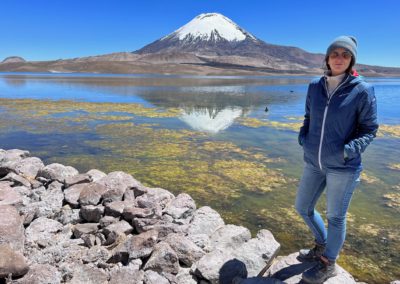 2022-11-14 South America Chile chilean Altiplano high plain plateau Andean Plateau Andes Mountains Andean Mountain Range Lauca National Park Lago Chungara lake mountains volcano snow Parinacota reflections in the water stones person people woman Ortovox sunglasses